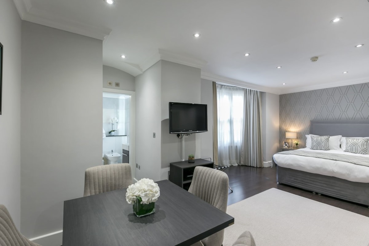 A modern hotel room with a bed, a TV, a dining table with chairs, and a bathroom visible in the background.