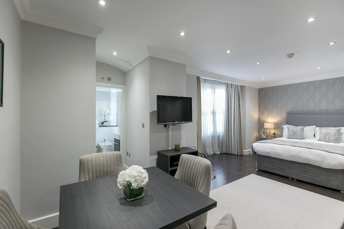 A modern hotel room with a large bed, a flat-screen TV, a dining area with a table and chairs, and a bathroom visible through an open door.