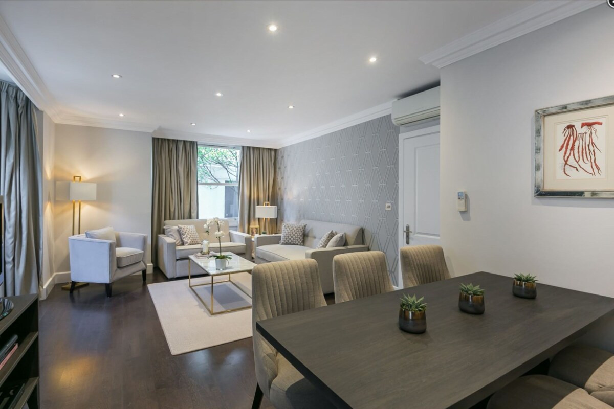 A modern living room with a sofa set, a dining table with potted plants, and a large window with curtains.