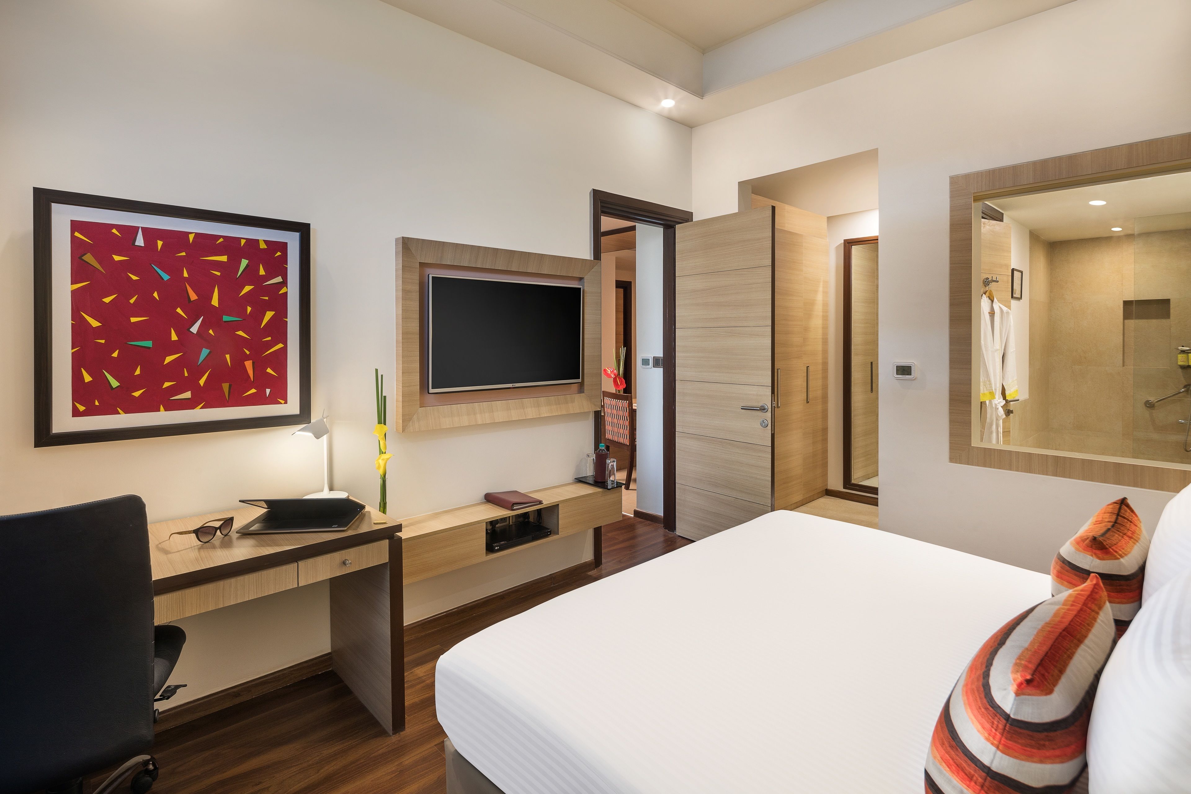 A modern hotel room with a bed, a desk with a computer, a TV, and a large mirror.