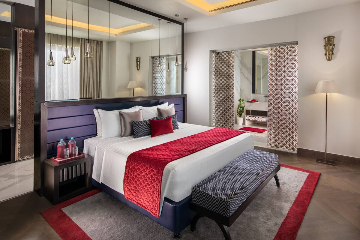 A modern hotel room with a large bed, bedside tables, a bench, a mirror, and a bathroom visible through a glass door.