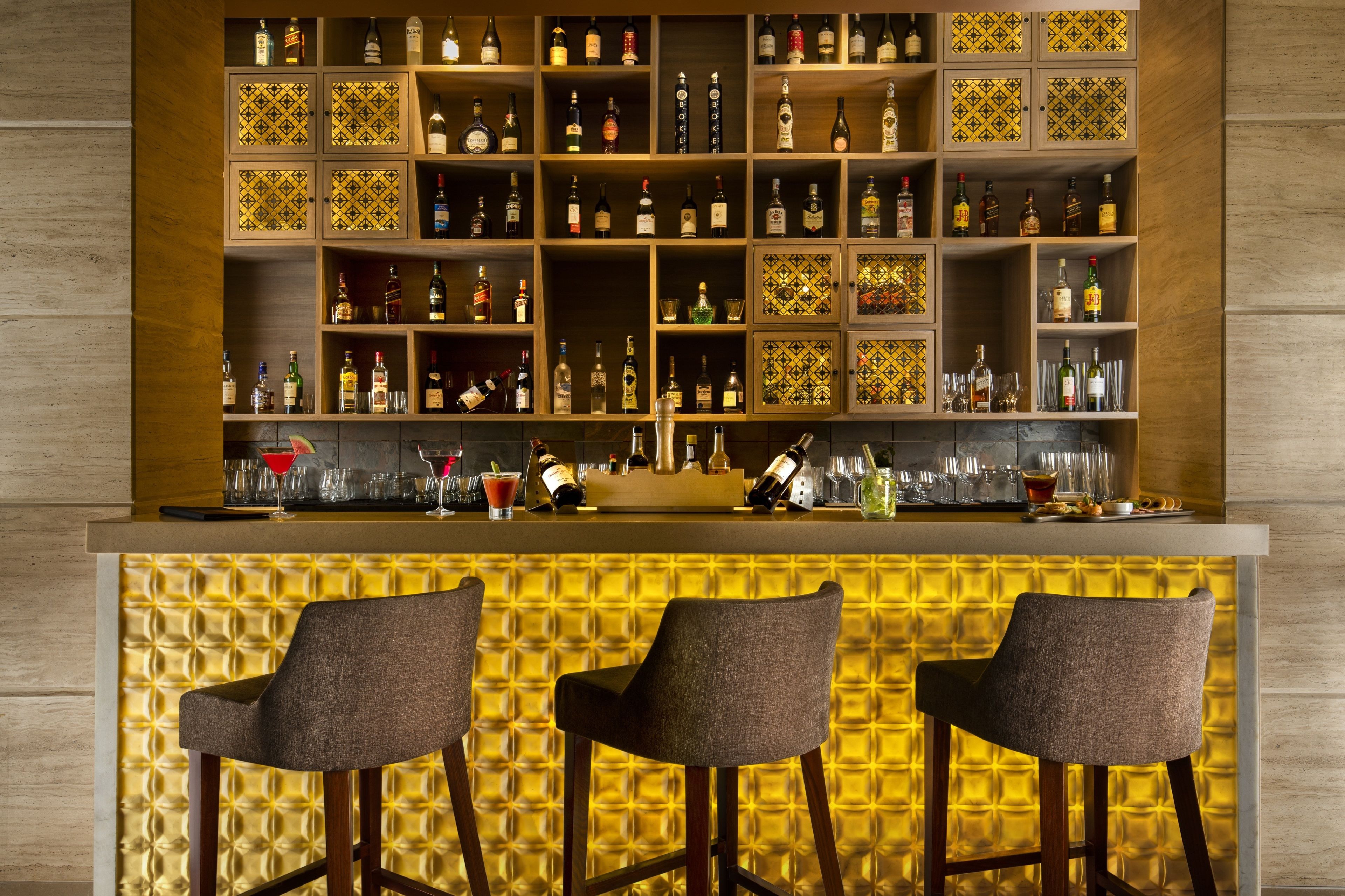 Zona de bar con varios estantes llenos de botellas, vasos y utensilios. Hay tres taburetes frente al mostrador.