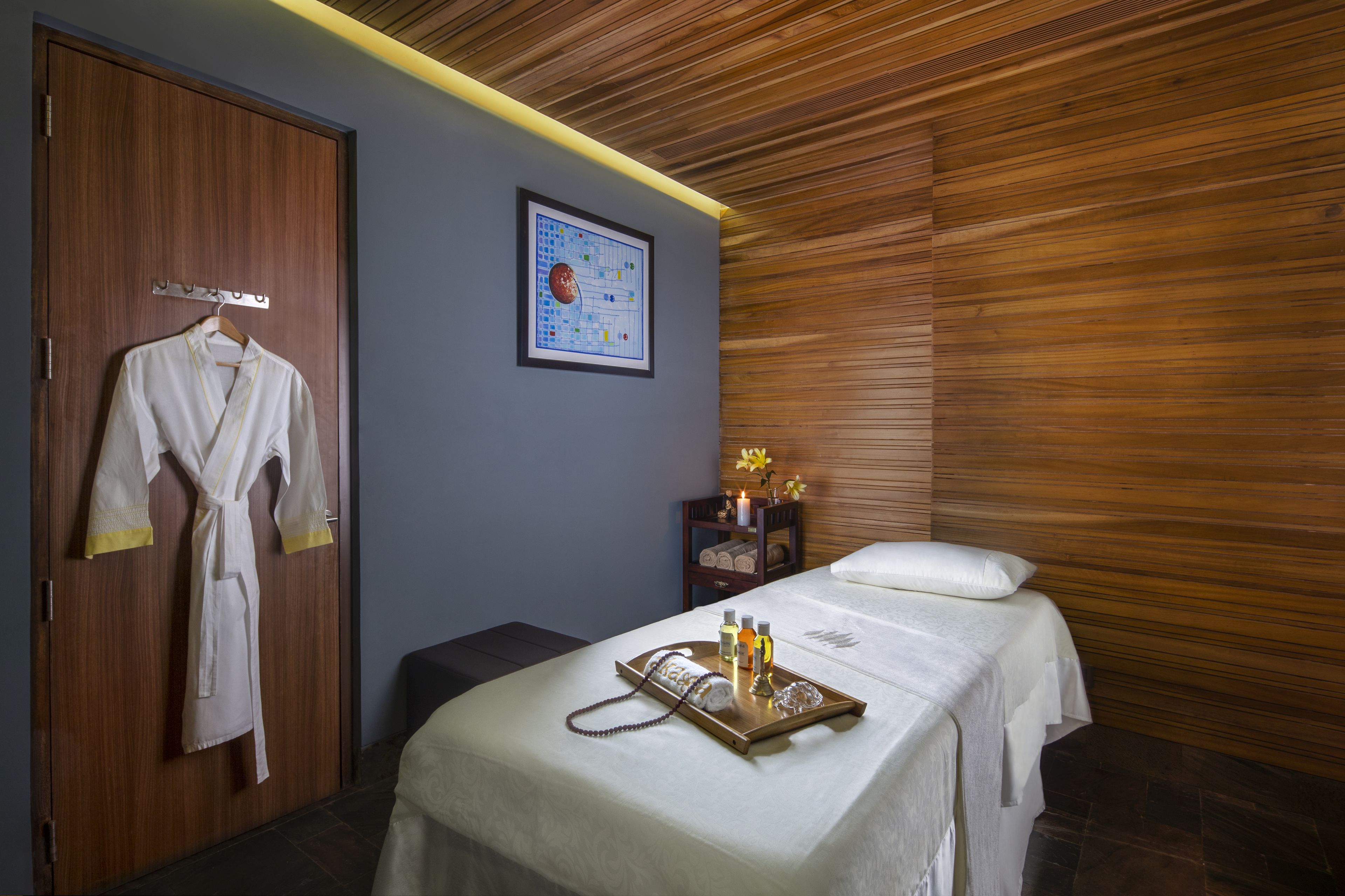 A room with a wooden ceiling, a bed with a tray of toiletries, a robe hanging on a door, and a framed picture on the wall.