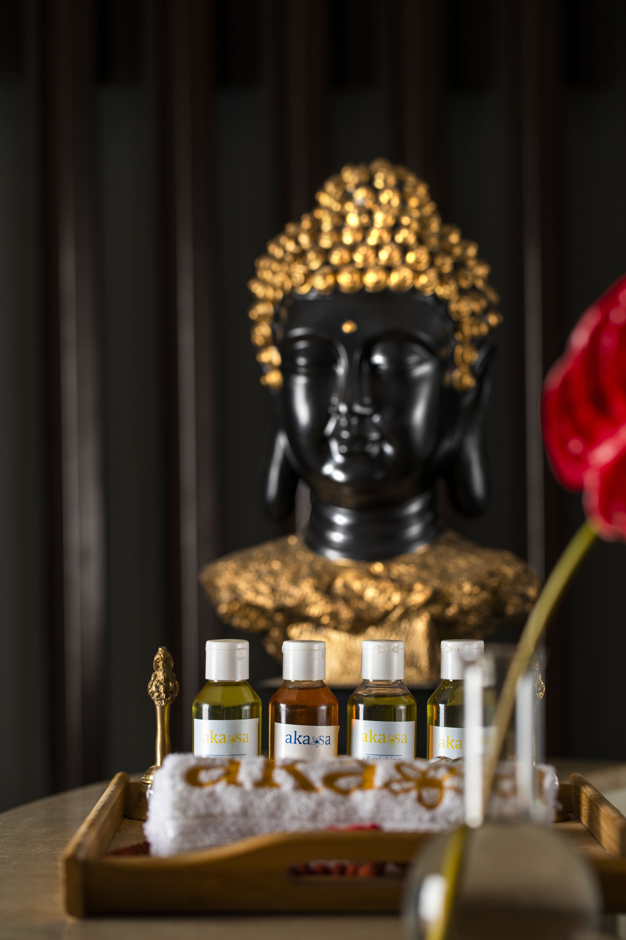 Une statue de Bouddha à la tête dorée, entourée de bouteilles étiquetées « akas », d’une serviette blanche et d’une rose rouge.