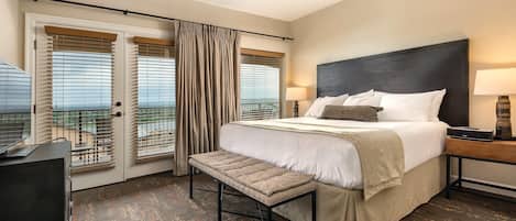 A hotel room with a large bed, a ceiling fan, a bench, and a view of the outdoors through the windows.