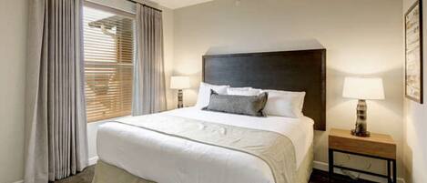 Una habitación de hotel con una cama grande, dos lámparas de noche, una mesita de noche de madera y una ventana con cortinas.