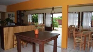 A dining area with a wooden table and chairs, a kitchen area with a counter, and a view of the outdoors through large windows. - Alecrim Pousada (Garopaba)