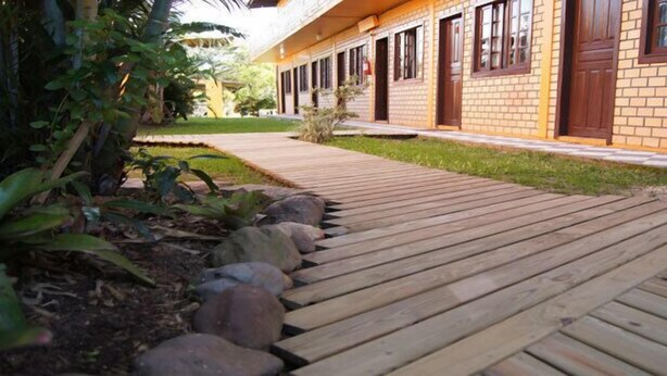 A wooden pathway leading to a brick building with multiple windows and doors. - Alecrim Pousada (Garopaba)