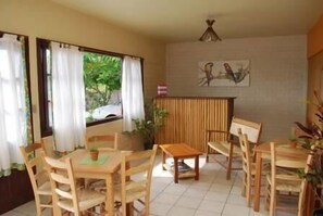A dining area with wooden furniture, a small table, and chairs. There are windows with curtains, a plant, and a wall-mounted artwork. - Alecrim Pousada (Garopaba)