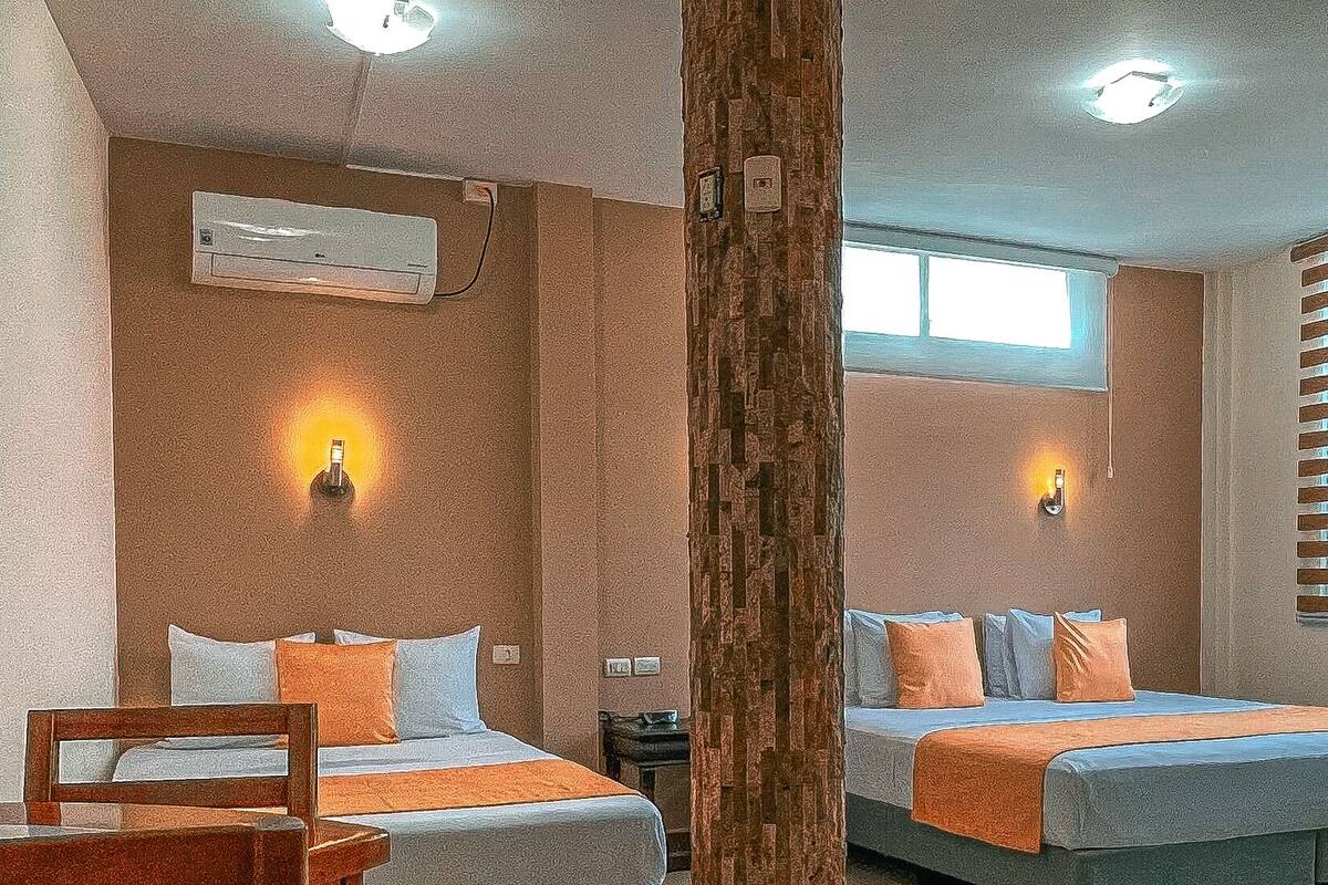 A hotel room with two beds, a wooden chair, a small table, a wooden column, and a window with blinds.
