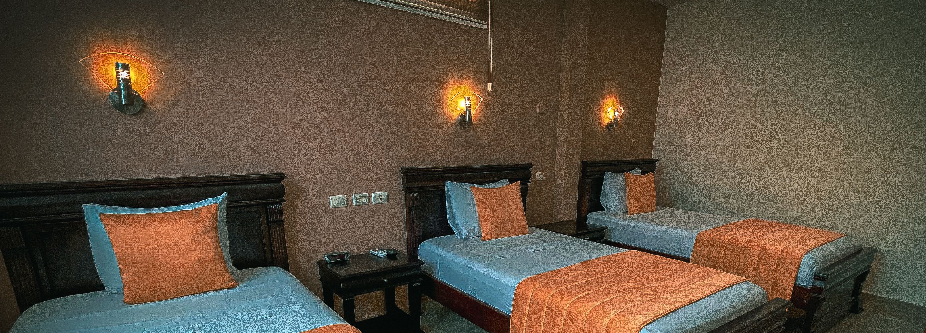 Una habitación de hotel con tres camas individuales, cada una con cabecero de madera y colcha azul. Hay dos apliques en las paredes a cada lado de la habitación.