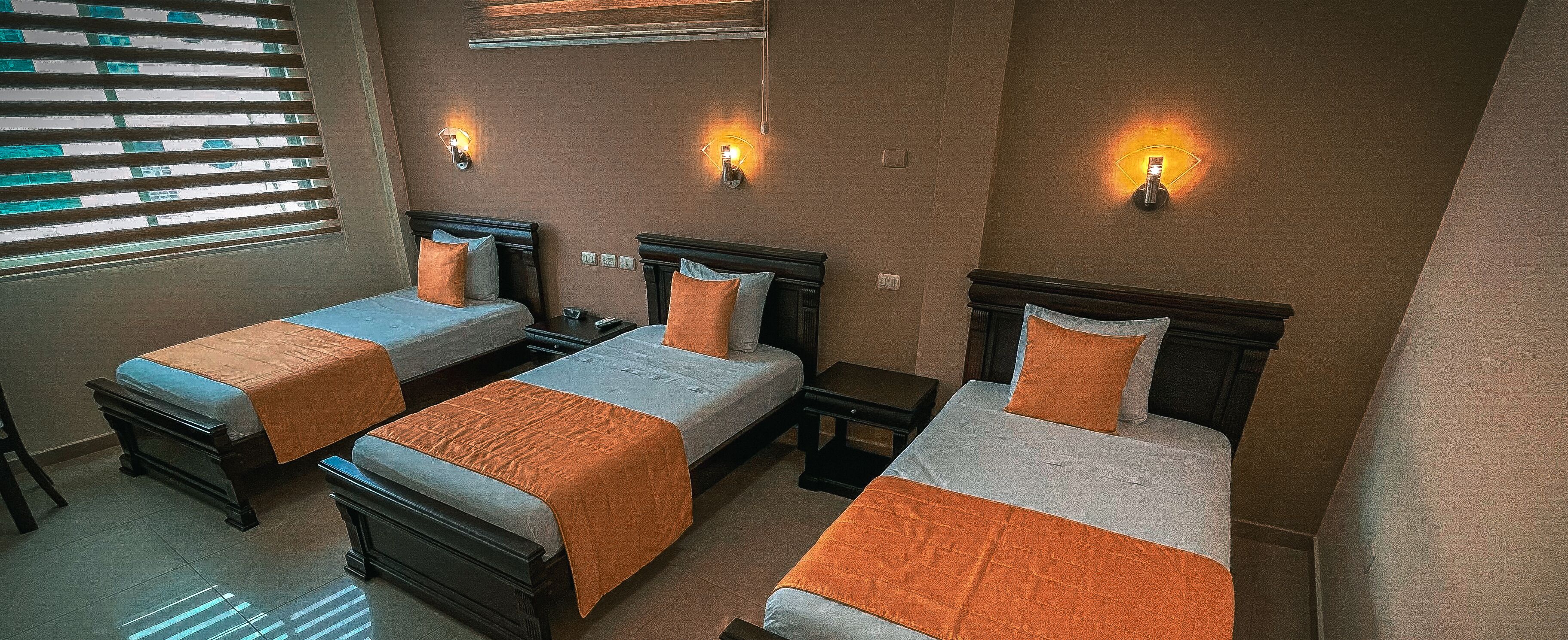 Une chambre d’hôtel avec trois lits simples, chacun recouvert d’une courtepointe orange et blanche, et dotée d’une tête de lit en bois. Des lampes murales sont fixées au-dessus de chaque lit.