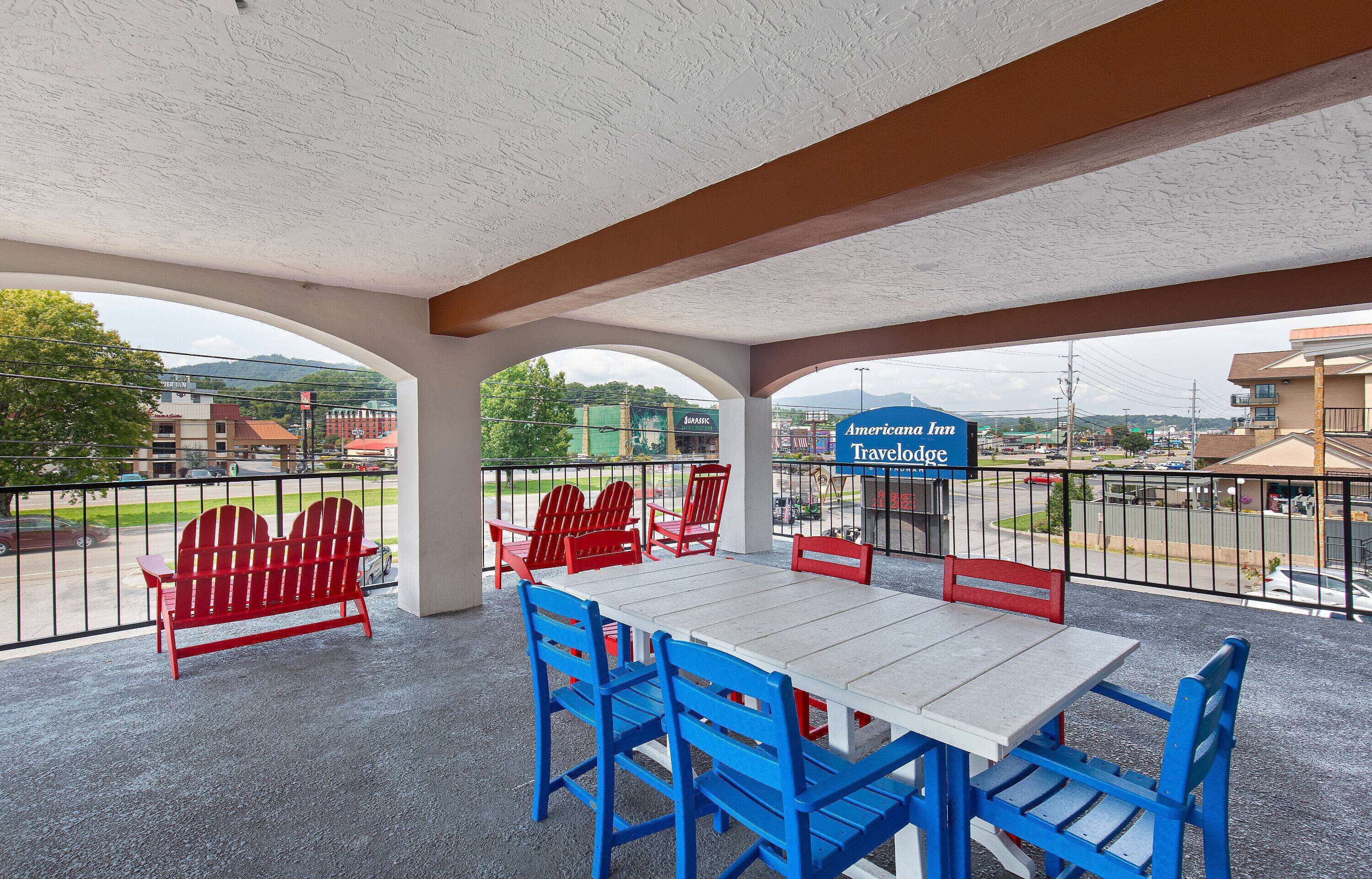 A covered outdoor seating area with a table and chairs, a sign for American Inn Travelodge, and a view of the parking lot and buildings in the background.