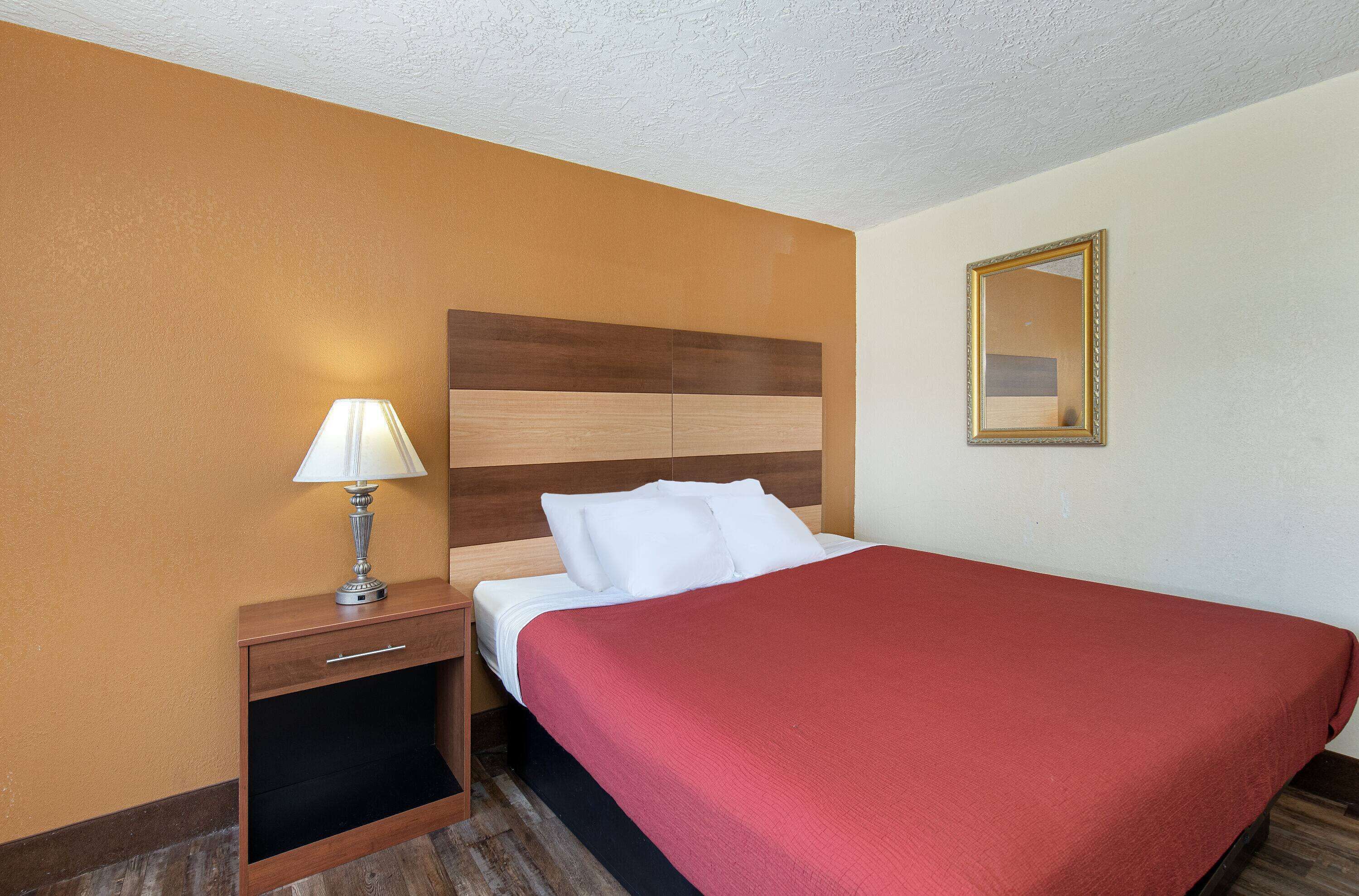 A hotel room with a wooden headboard, a bed with a red bedspread, a nightstand with a lamp, and a mirror on the wall.