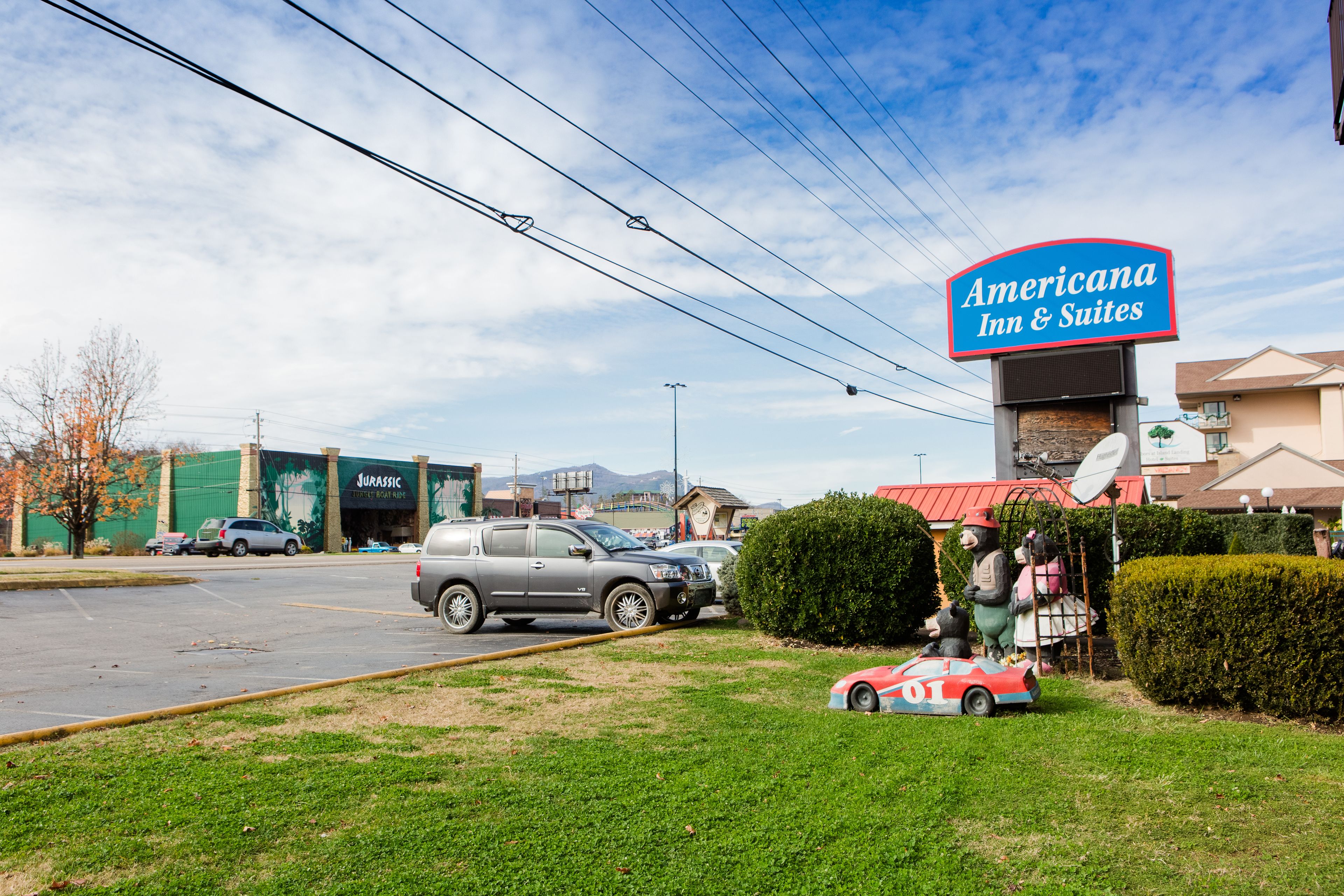 A parking lot with a sign for Americana Inn & Suites, a Jurassic-themed building, and a statue of a bear with a car.