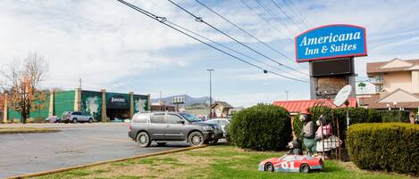 A parking lot with a sign for Americana Inn & Suites, a Jurassic-themed building, and a statue of a bear with a car.