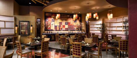 A restaurant interior with wooden furniture, a central bar, and a decorative wall with a geometric pattern.