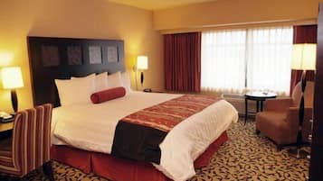 A hotel room with a large bed, a chair, a small table, and a window with curtains.