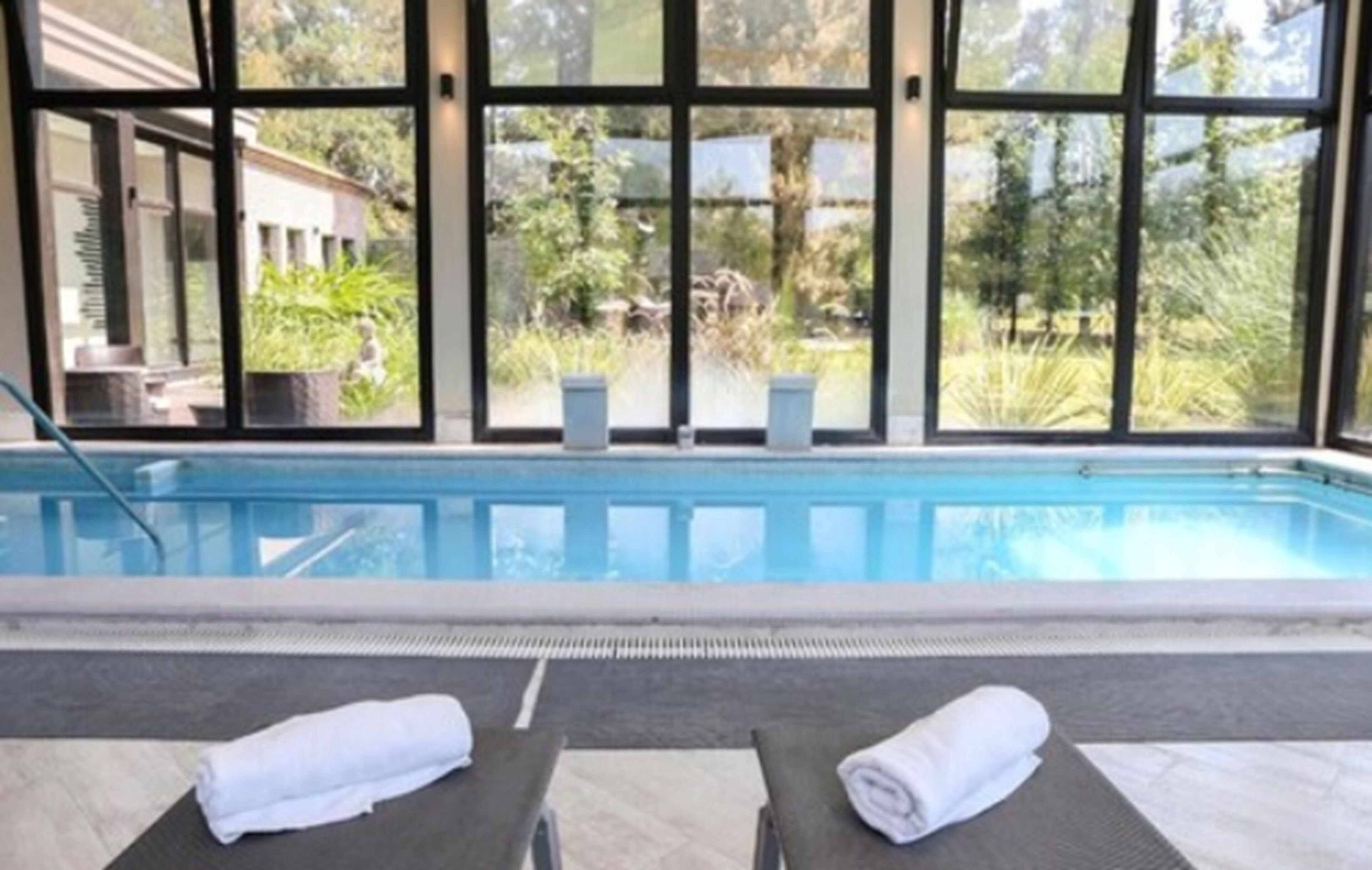 Zona de piscina con dos sillas de descanso y vistas a un edificio y follaje a través de amplios ventanales.