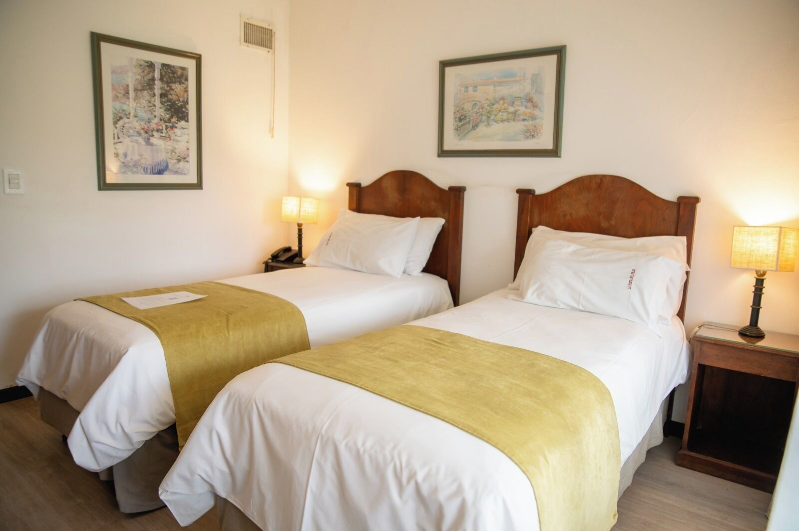 Een hotelkamer met twee enkele bedden, elk met een houten hoofdbord, wit beddengoed en een gele plaid. Er zijn twee nachtkastjes met lampen, en de muren zijn versierd met ingelijste kunstwerken.