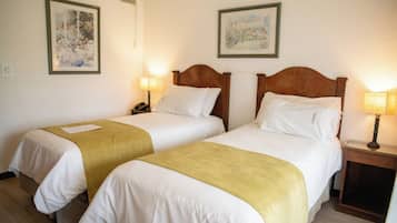 Een hotelkamer met twee enkele bedden, elk met een houten hoofdbord, wit beddengoed en een gele plaid. Er zijn twee nachtkastjes met lampen, en de muren zijn versierd met ingelijste kunstwerken.