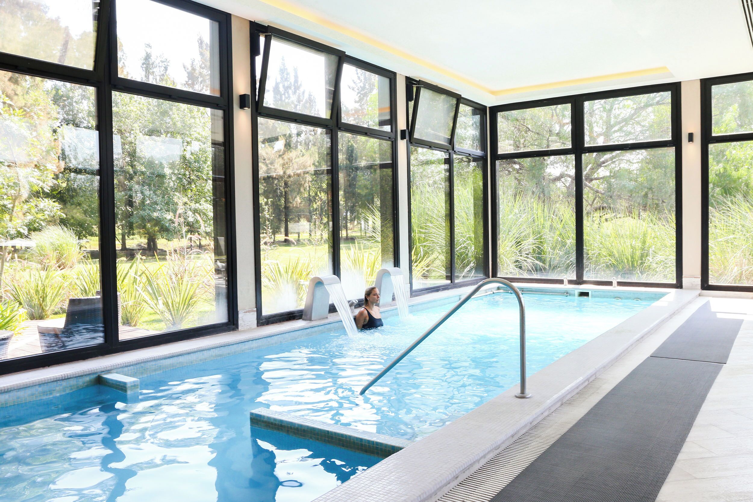 An indoor swimming pool with clear glass walls, a ladder, and a view of greenery outside.