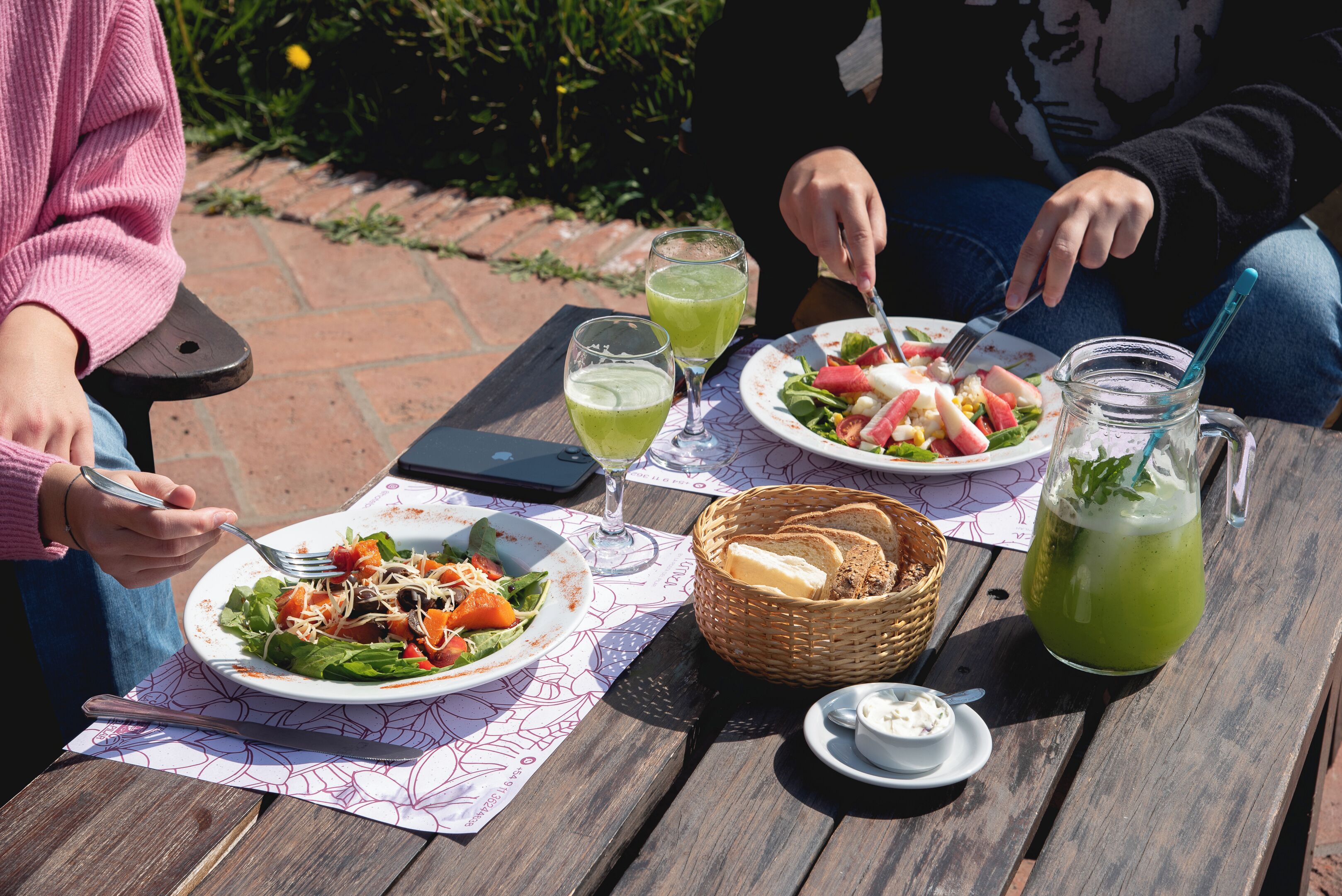 Une table en bois sur laquelle est disposé un repas composé d’une salade, de pain et d’une boisson verte, ainsi qu’un panier et un petit récipient contenant de la crème.