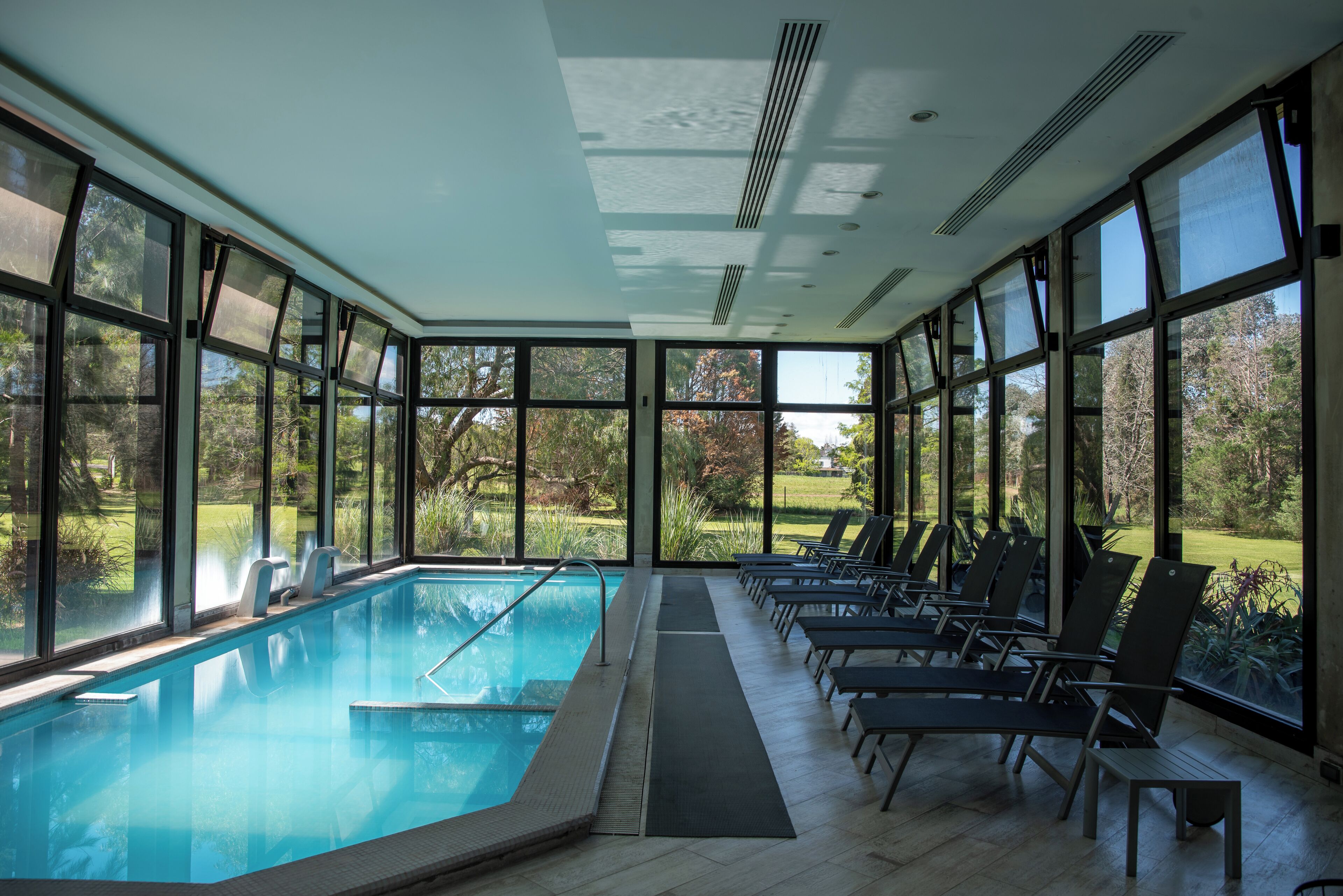 An indoor swimming pool with clear views of the outdoors, surrounded by large windows and equipped with lounge chairs.
