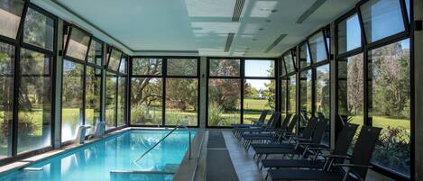 An indoor swimming pool with clear views of the outdoors, surrounded by large windows and equipped with lounge chairs.