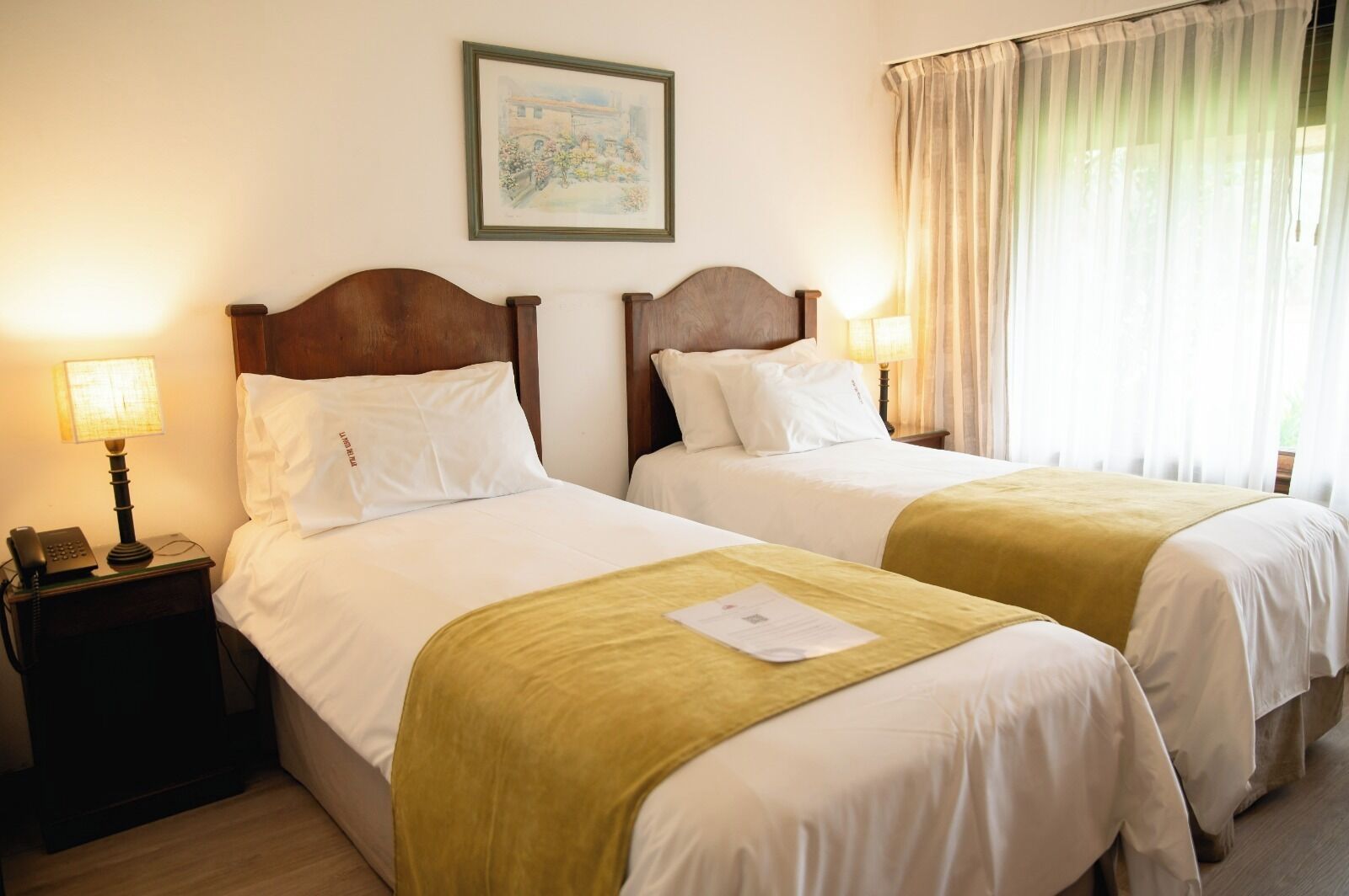 Habitación de hotel con dos camas, una mesita de noche con lámpara y un cuadro en la pared.