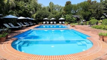 Una piscina rectangular con borde de ladrillo, rodeada de sillas de descanso y sombrillas, ubicada en un área ajardinada con árboles y arbustos.