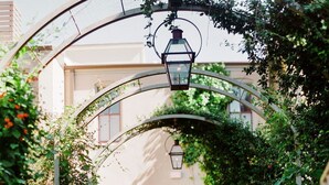 Un sendero en un jardín que lleva a un edificio con puerta roja, rodeado de abundante follaje y un arco de metal con un farol.