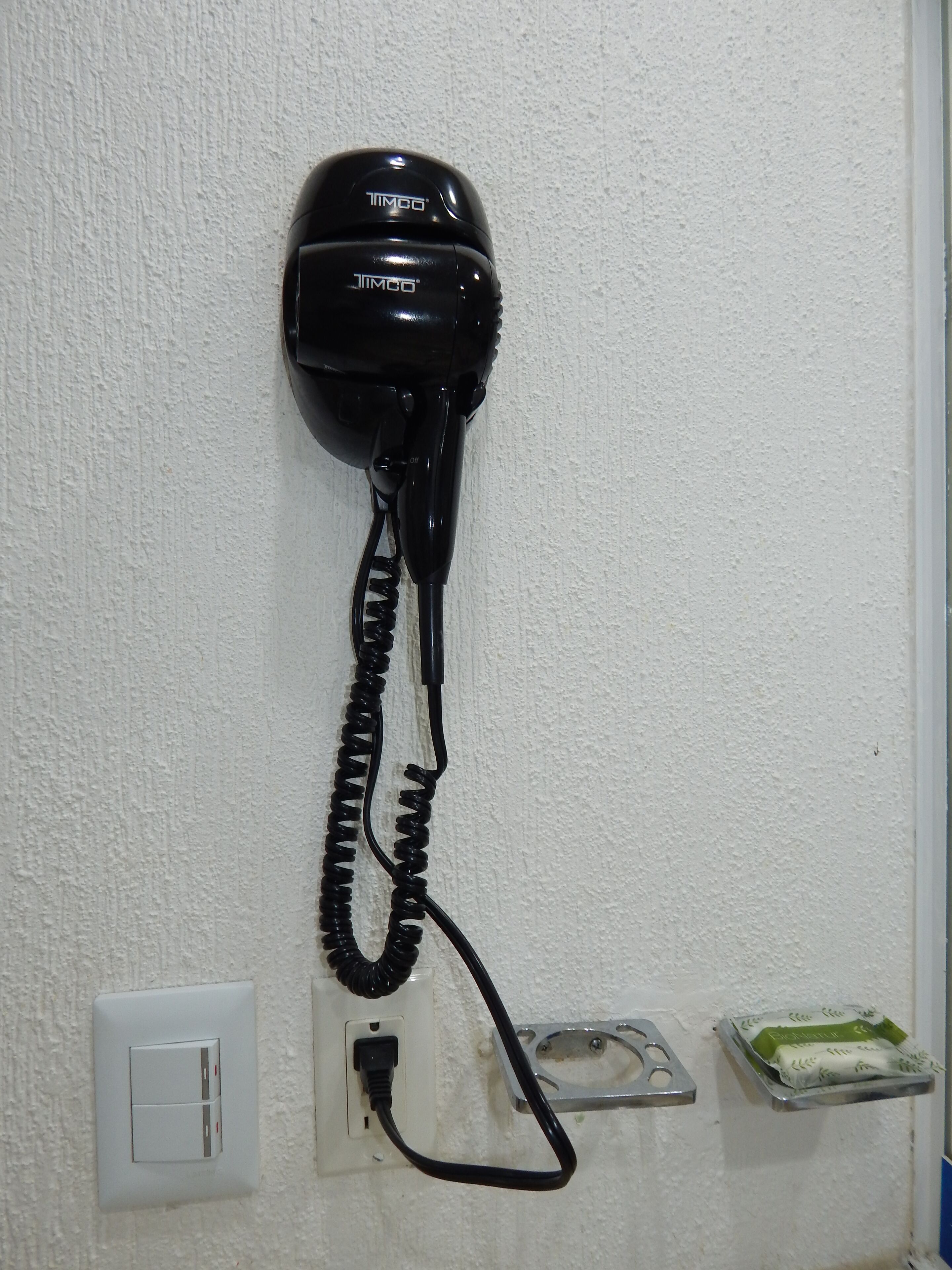 Un teléfono negro fijado en una pared blanca texturizada, conectado a una toma de corriente y una bandeja metálica con una caja de pañuelos.