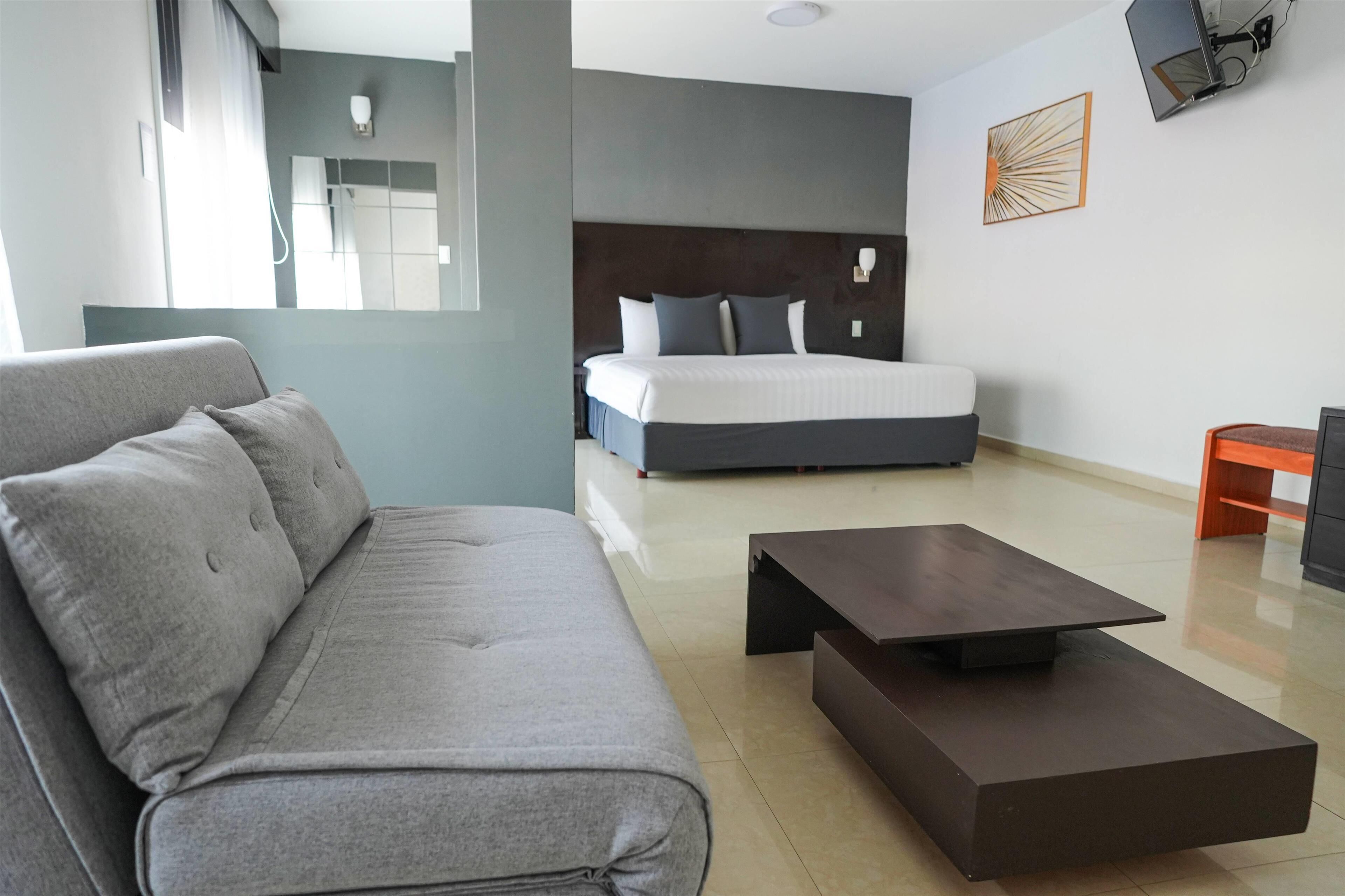 A modern hotel room with a grey sofa, a bed with a dark headboard, a small wooden side table, and a flat-screen TV mounted on the wall.