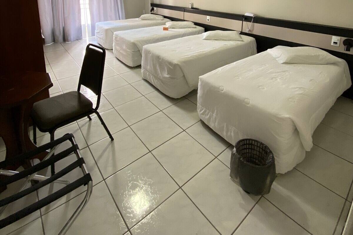 A hotel room with three single beds, a chair, a trash bin, and a window with curtains.