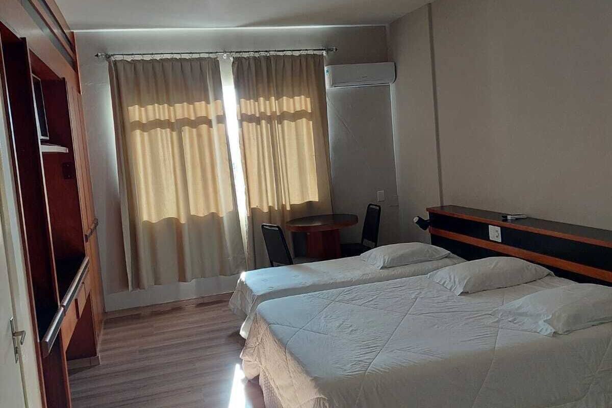 A hotel room with a wooden floor, two beds, a desk with chairs, and a window with curtains.