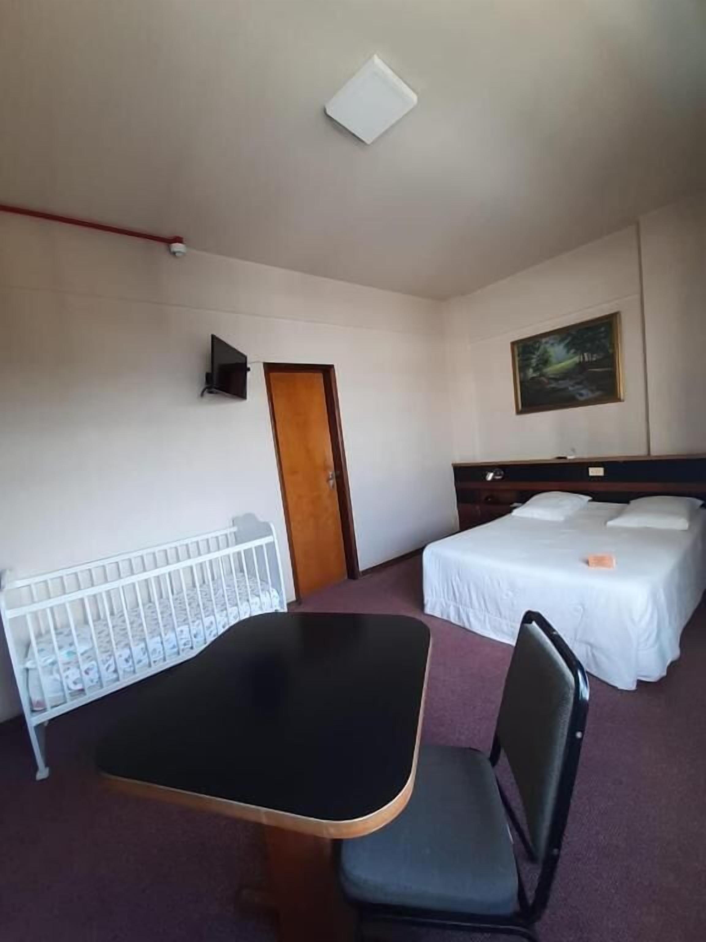 Quarto de hotel com berço, uma cama de solteiro, uma cadeira, uma mesa e um quadro na parede.