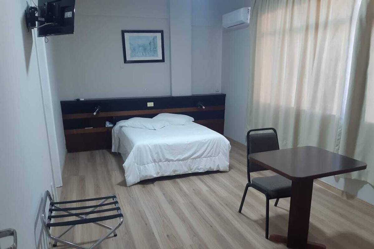 A hotel room with a bed, a table, and a chair. There is a window with curtains, a picture on the wall, and an air conditioning unit.