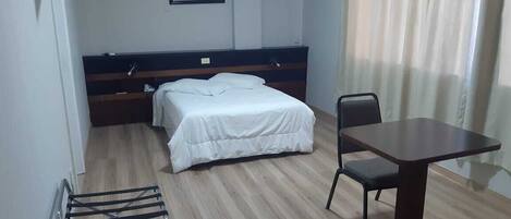 Ein Hotelzimmer mit einem Bett, einem Tisch und einem Sessel. Es gibt ein Fenster mit Vorhängen, ein Bild an der Wand und eine Klimaanlage.