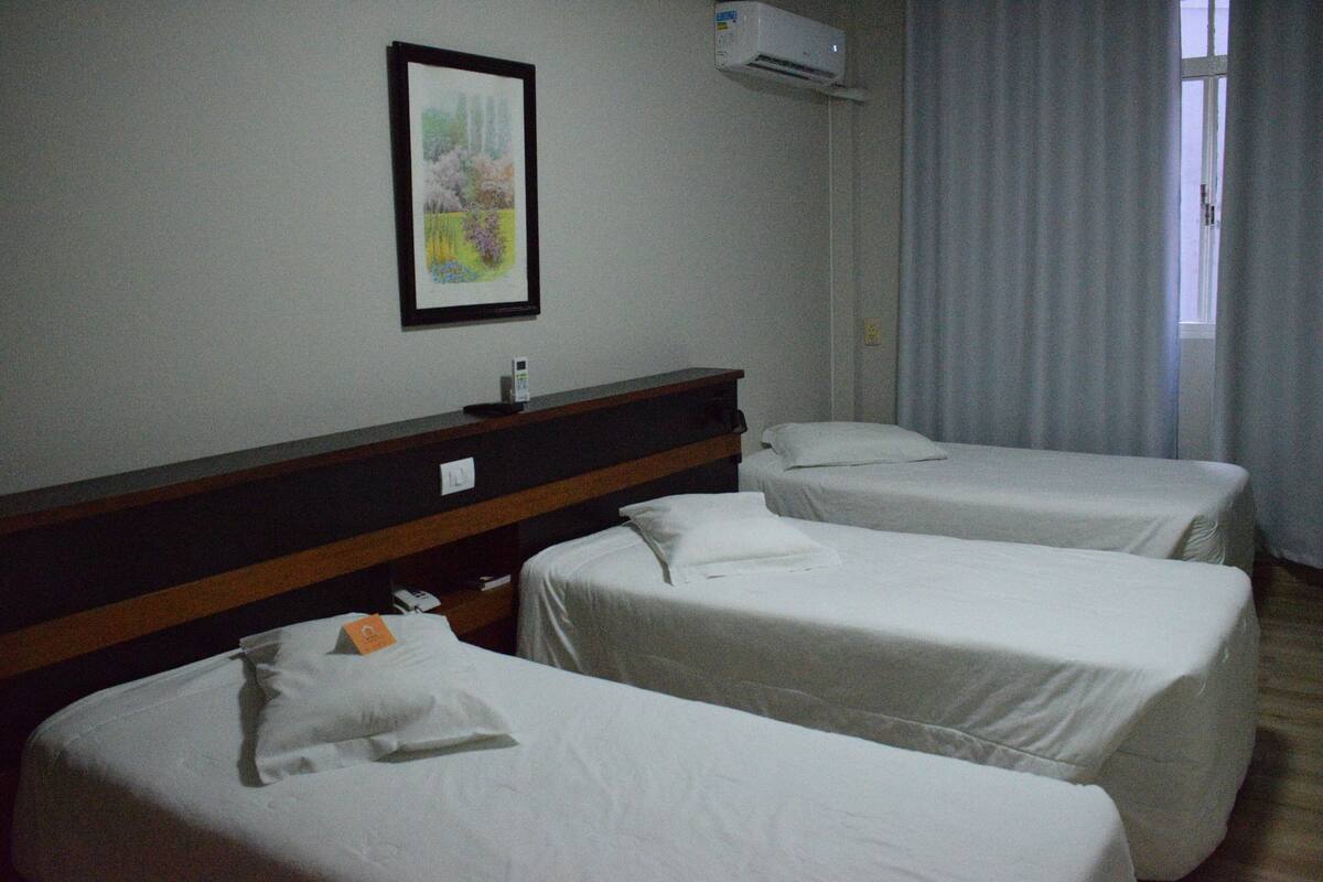 A hotel room with three single beds, a wooden headboard, a framed picture on the wall, and an air conditioning unit.
