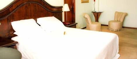 A neatly made bed with white linens, a wooden headboard, a bedside table with a lamp, and two chairs in the room.