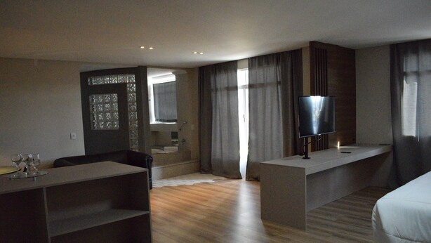 A modern hotel room with a wooden floor, a flat-screen TV on a stand, a bed, a desk with a chair, and a small seating area with a table and chairs.