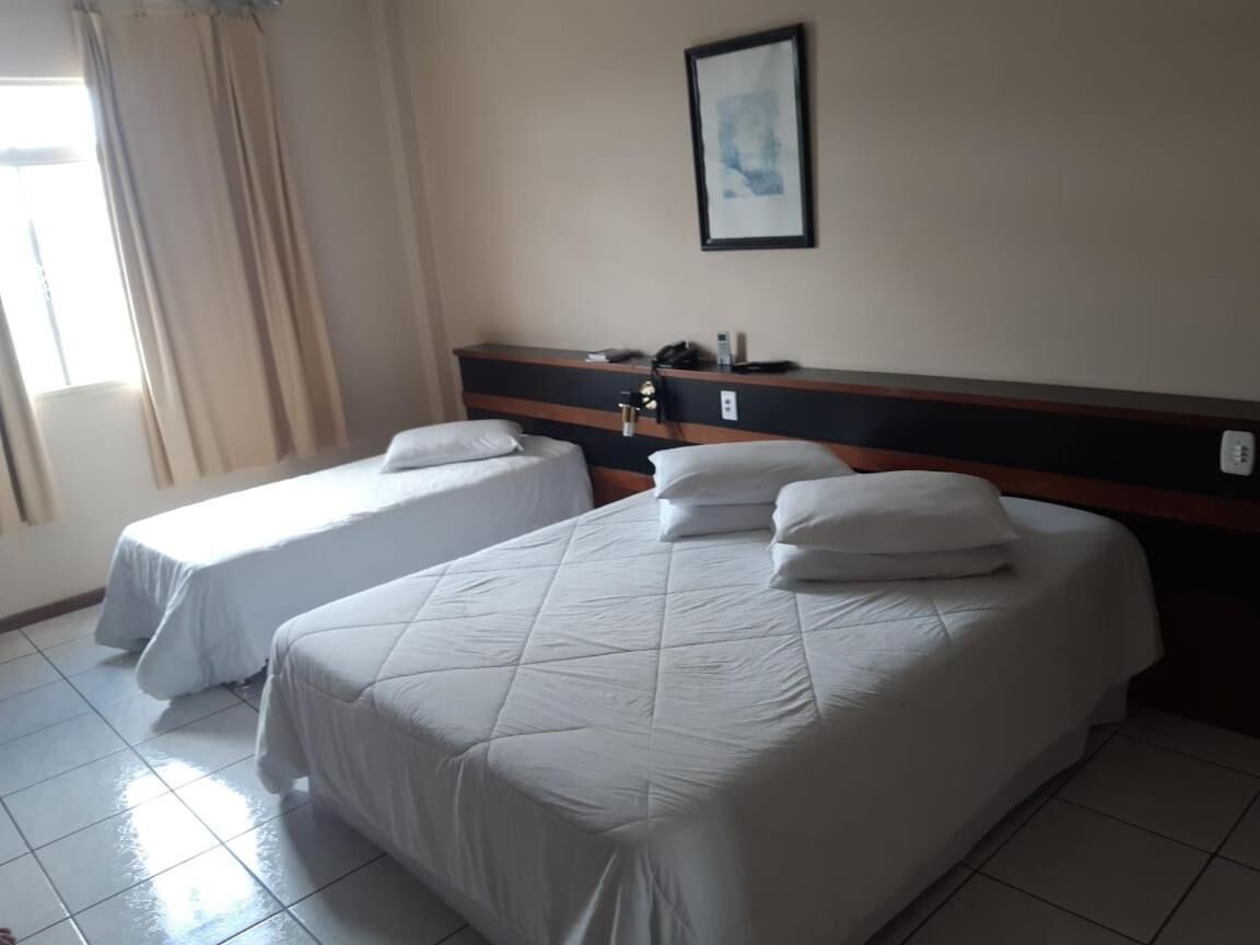 Quarto de hotel com duas camas, cabeceira, mesa de cabeceira, um quadro emoldurado na parede e uma janela com cortinas.