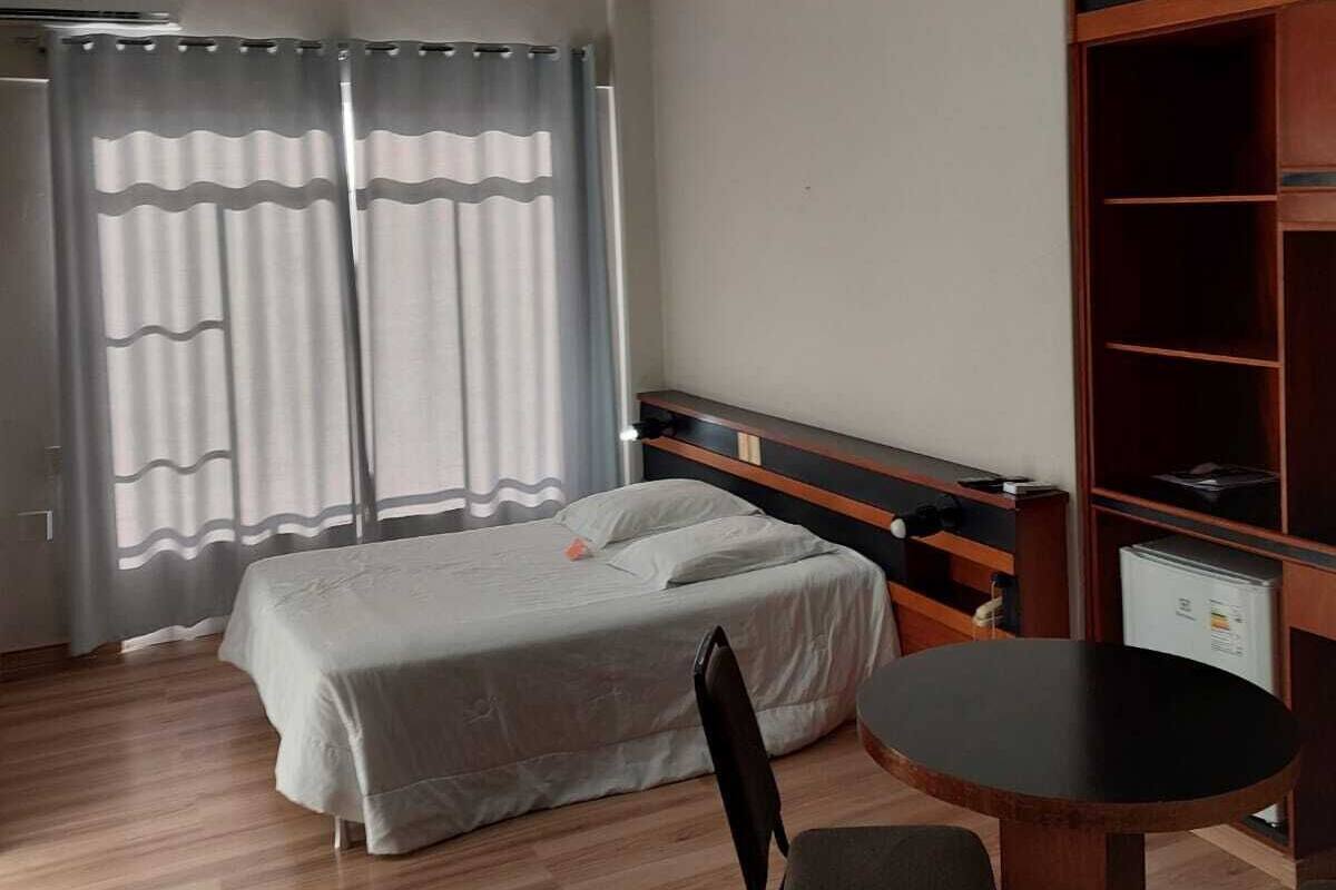 A hotel room with a bed, a desk, and a chair. There is a window with curtains, a ceiling light, and a wardrobe.