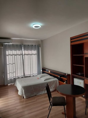 A hotel room with a bed, a desk, and a chair. There is a window with curtains, a ceiling light, and a wardrobe.