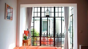 Ein Schlafzimmer mit großem Fenster, einem Bett mit bunten Kissen, einem kleinen Tisch mit Pflanze und einer Pendelleuchte.