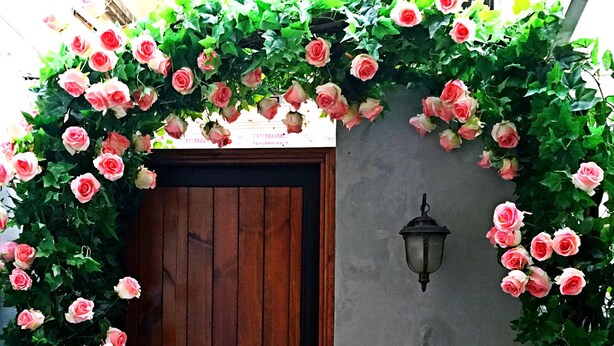 Eine Holztür, umrahmt von einem üppigen Rosenbogen in Rosa und Weiß, mit einer an der Wand befestigten Leuchte rechts davon.