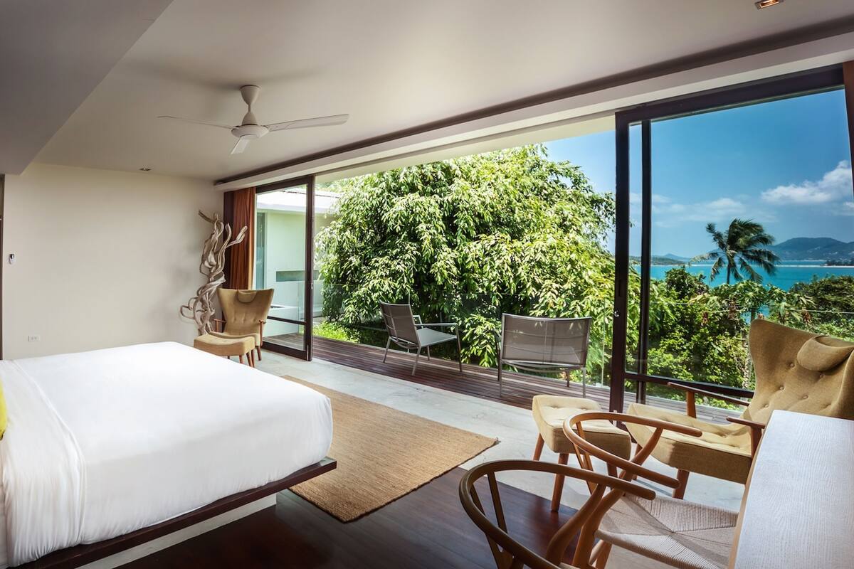 A modern hotel room with a large bed, a balcony with seating, and a view of the ocean and trees.