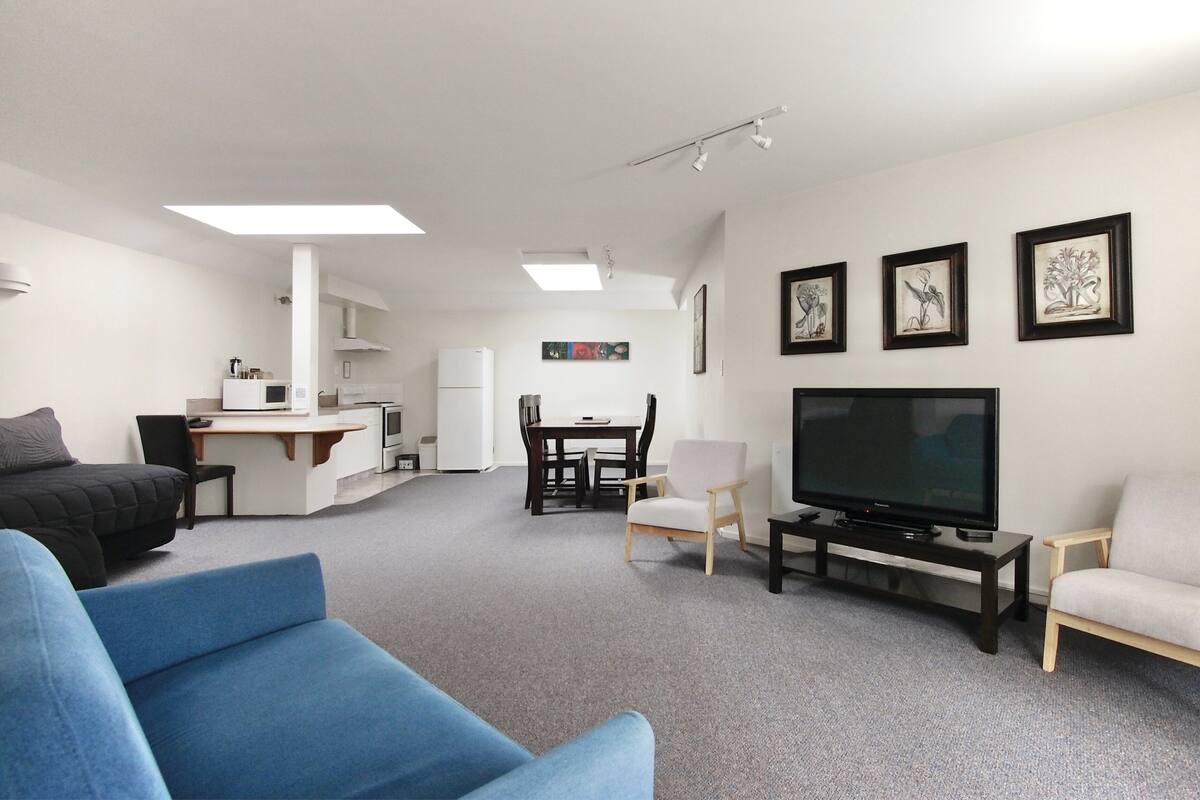 A modern living room with a flat-screen TV, a dining area, and a kitchen in the background.
