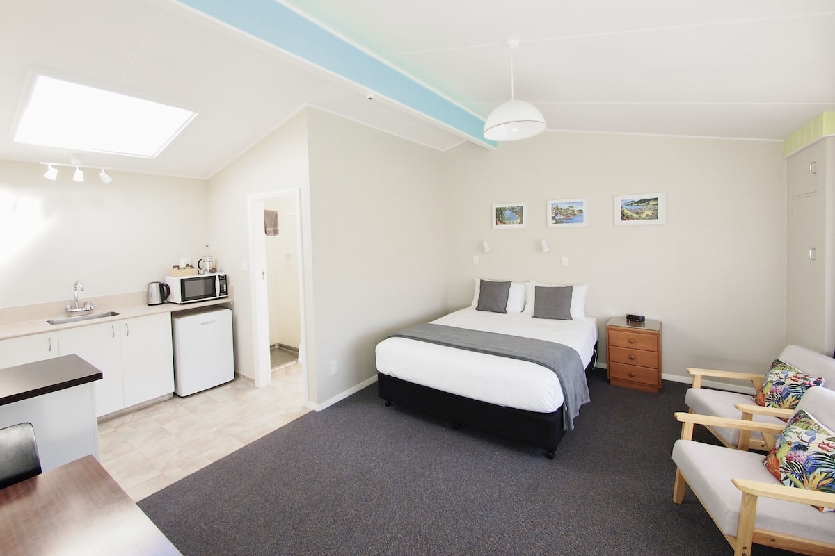 A bedroom with a bed, bedside table, and a desk with a chair. There is a kitchenette with a microwave and a sink.